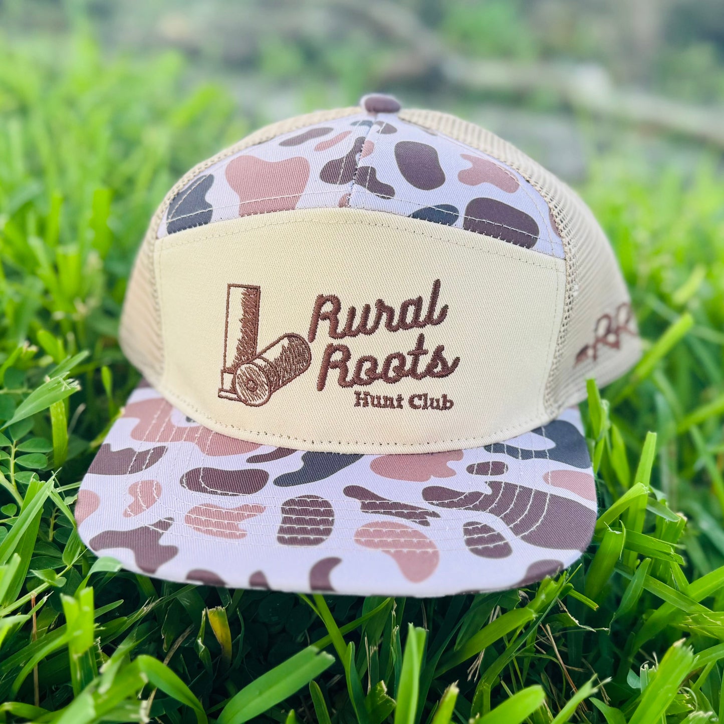 Trucker hat with camouflage pattern and 'Rural Roots' logo on grass